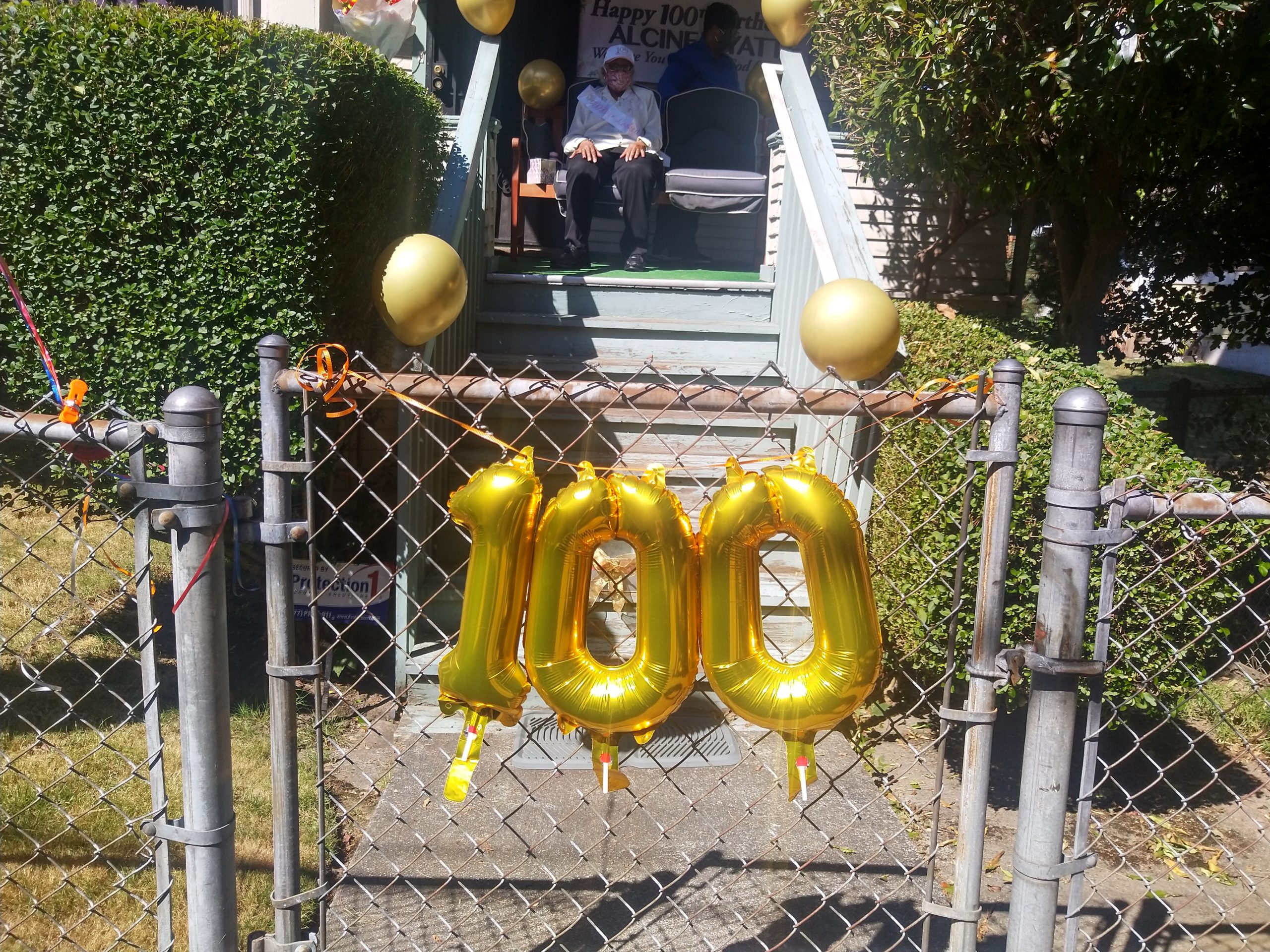 Image description: A chain link fence decorate with balloons in the shape of the number 100. Behind the fence is a porch, and on the porch sits an elderly woman wearing a birthday sash.