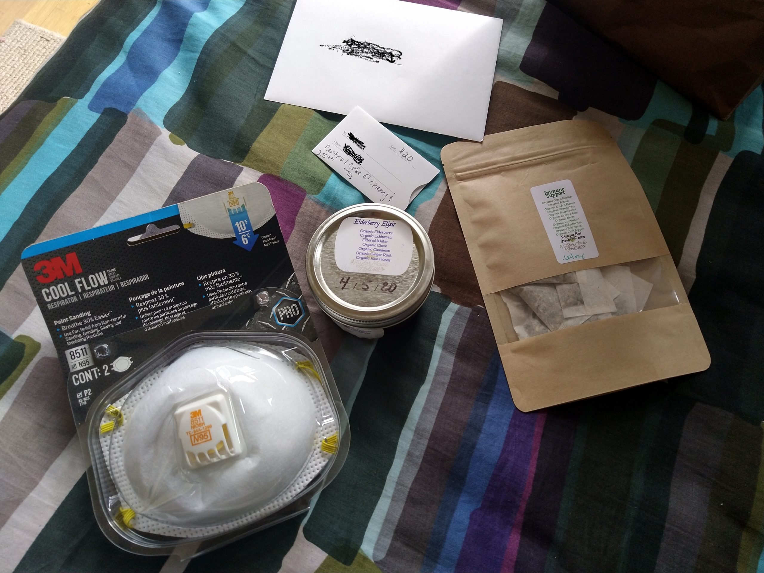 Image descriptions: Several items displaced on a purple, blue, and green bedspread: a package of N95 masks, a jar of elderberry syrup, a sealed bag of immune tea, and a $20 gift card